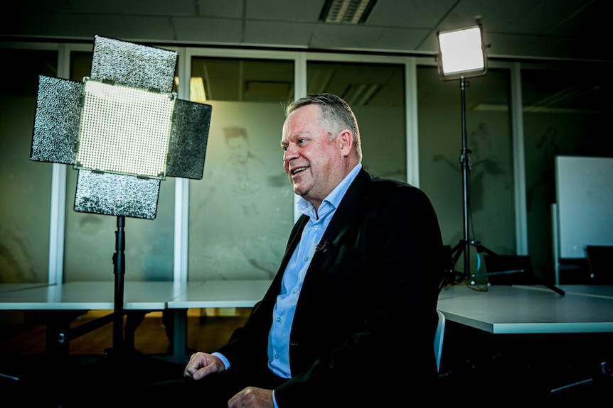 A man in a suit sits among lighting for television cameras.