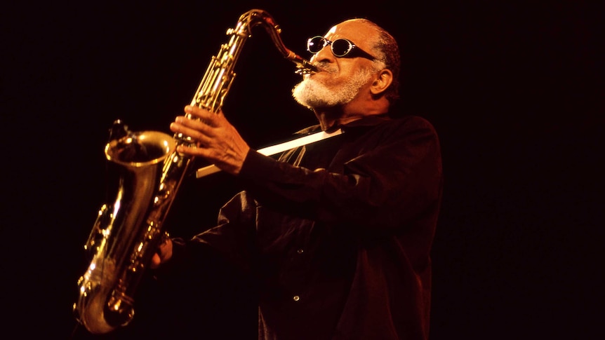 Against a dark background, a man wearing glasses with short white beard plays a saxophone, holding it up high and tensing face.
