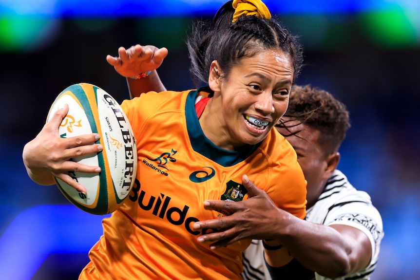 A Wallaroos player holds the ball with her right hand as she is tackled by a Fijiana opponent.