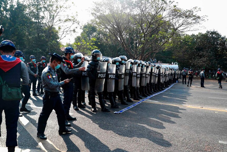 A line of riot police stand shoulder to shoulder with their shields raised.
