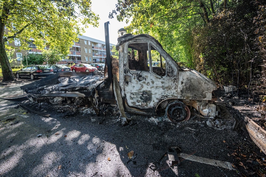 A burnt out truck is seen in the street with trees in the background.