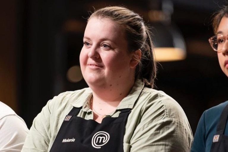 Melanie Persson, wearing black apron with Masterchef 'm' logo, stands smiling with closed mouth looking ahead.