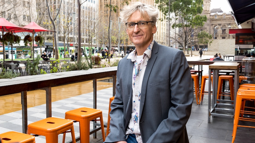 A man with wavy grey hair, wearing a jacket and glasses, sitting at an open-air cafe in Melbourne.