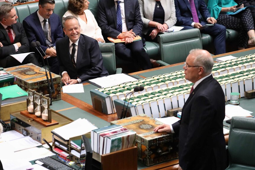 Mr Shorten is watching Mr Morrison and smiling during Question Time. They are on opposite sides of the despatch boxes.