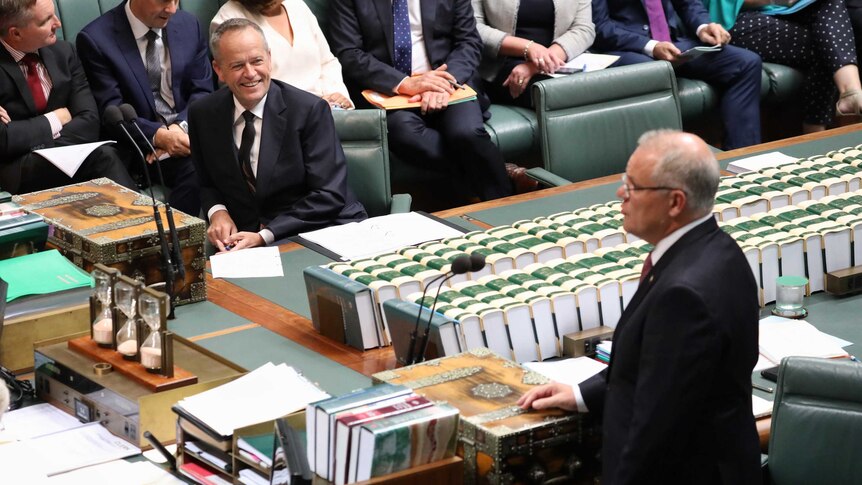 Mr Shorten is watching Mr Morrison and smiling during Question Time. They are on opposite sides of the despatch boxes.