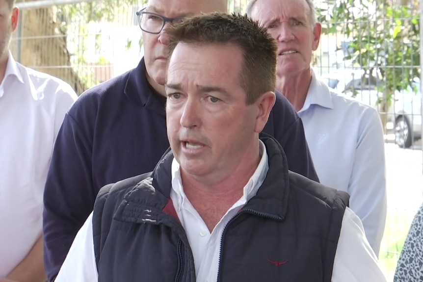 Man in white shirt and black zip vest talking, several people standing behind him