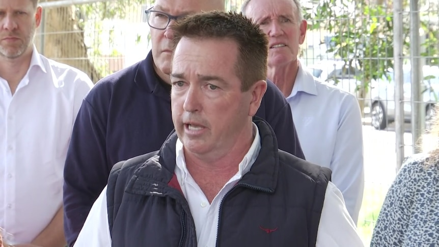 Man in white shirt and black zip vest talking, several people standing behind him