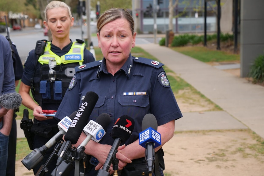 A middle-aged woman in a police uniform stands outdoors and speaks to the media.