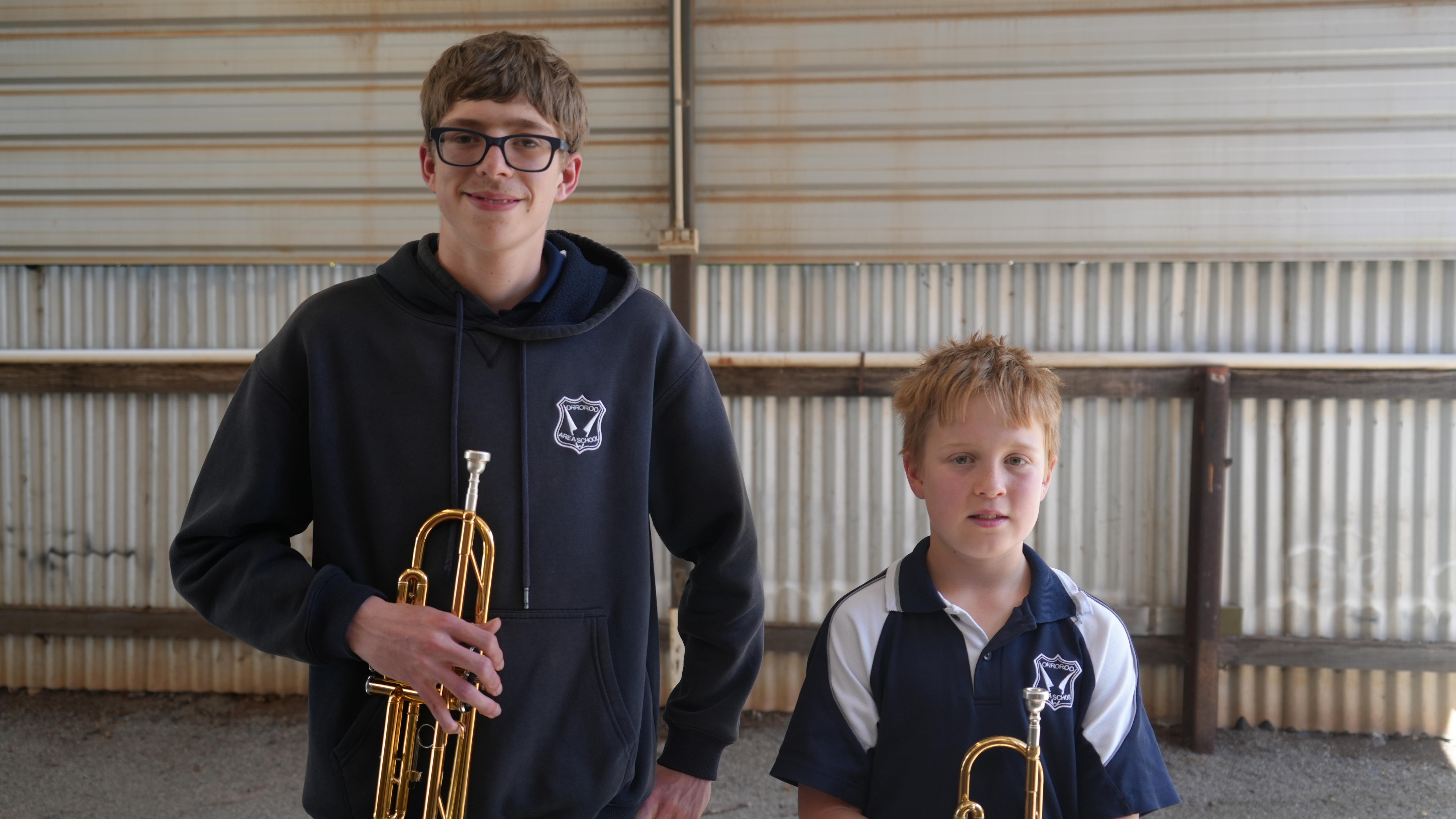 A tall teenage boy and a short young boy, both holding trumpets and smiling at the camera.
