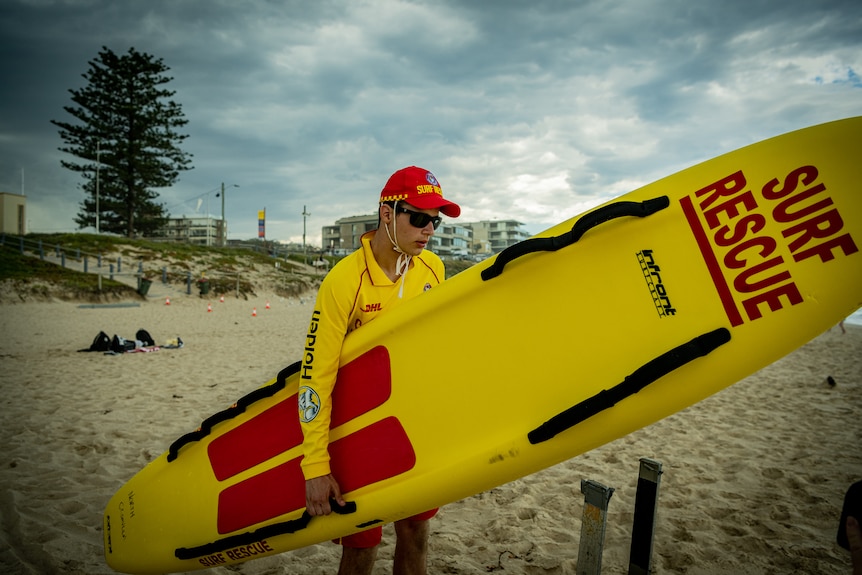 Syros carries a large yellow surf rescue longboard down the beach.