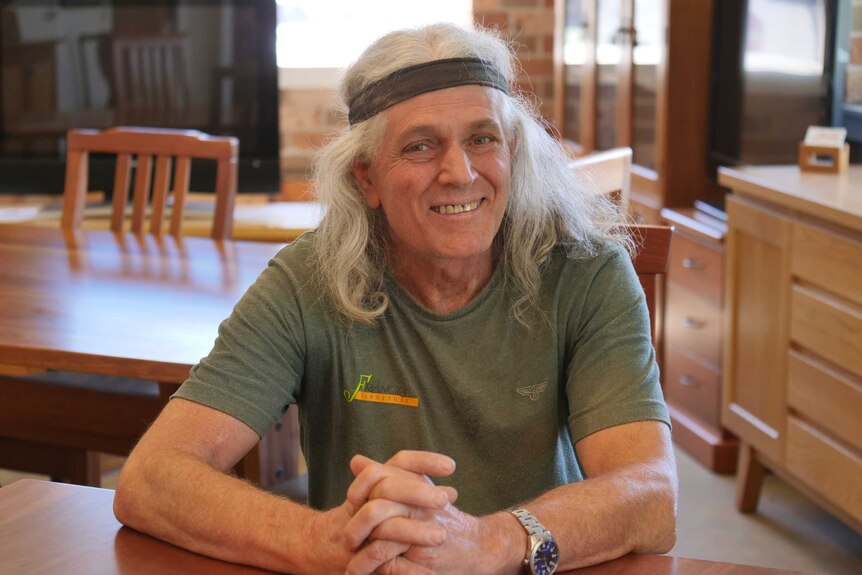 A man with long grey hair sits at a timber table smiling.