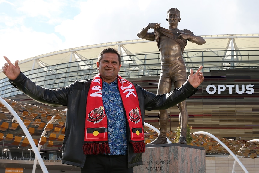 Nicky Winmar stands with outstretched arms in front of a statue at Perth Stadium of his iconic stand against racism.