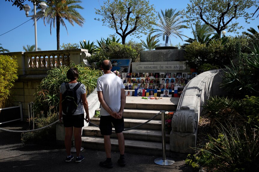 Two people pictured from behind looking at a memorial in Nice with photos of victims and 'In Memory of our Angels' engraved