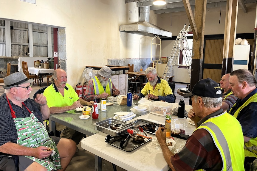 A group of men, most wearing high visibility work wear and vests sit around pushed-together tables enjoying a barbecue lunch