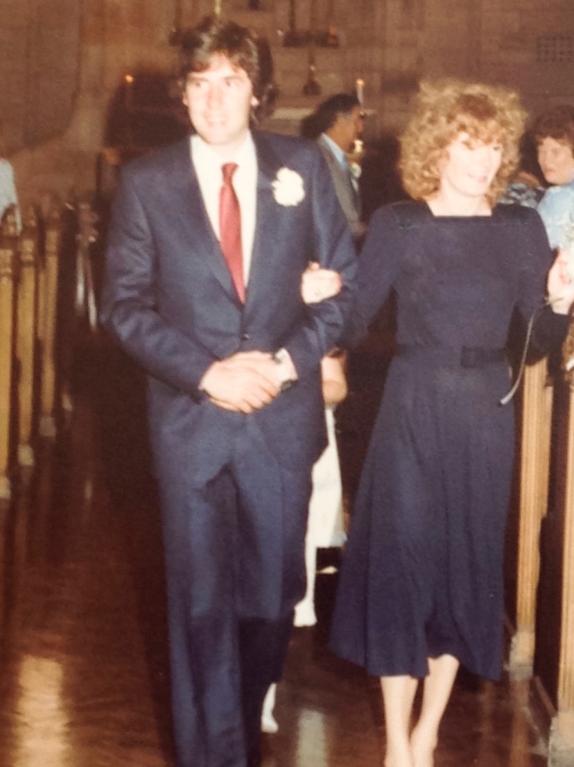 A newly married couple walking down the aisle at a chapel, with the bride wearing a navy dress.