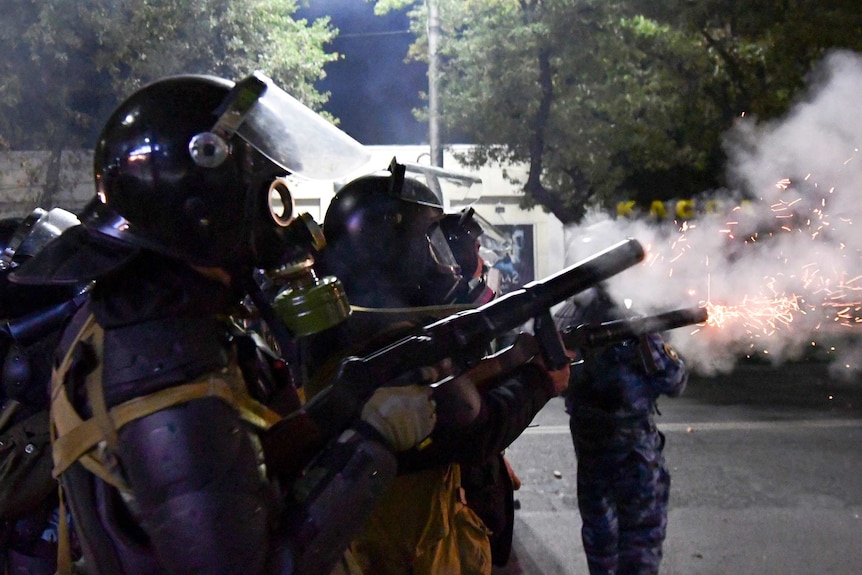 Riot police wearing helmets and gas masks stand in a line and fire tear gas