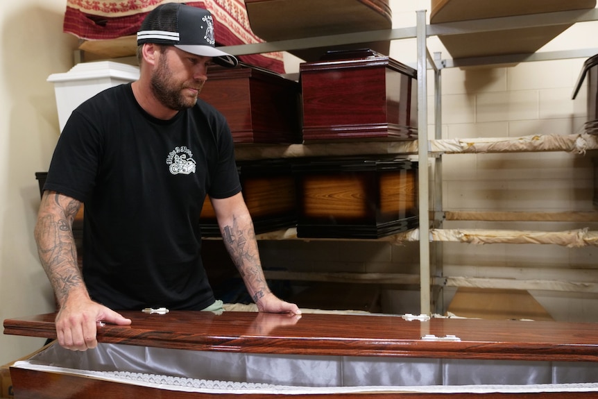 A man with facial hair and tattoos looks at a casket. There are stacks of caskets in the background