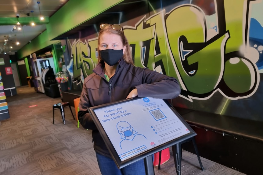 A woman standing behind a COVID-19 check in sign with spray painted walls behind her.