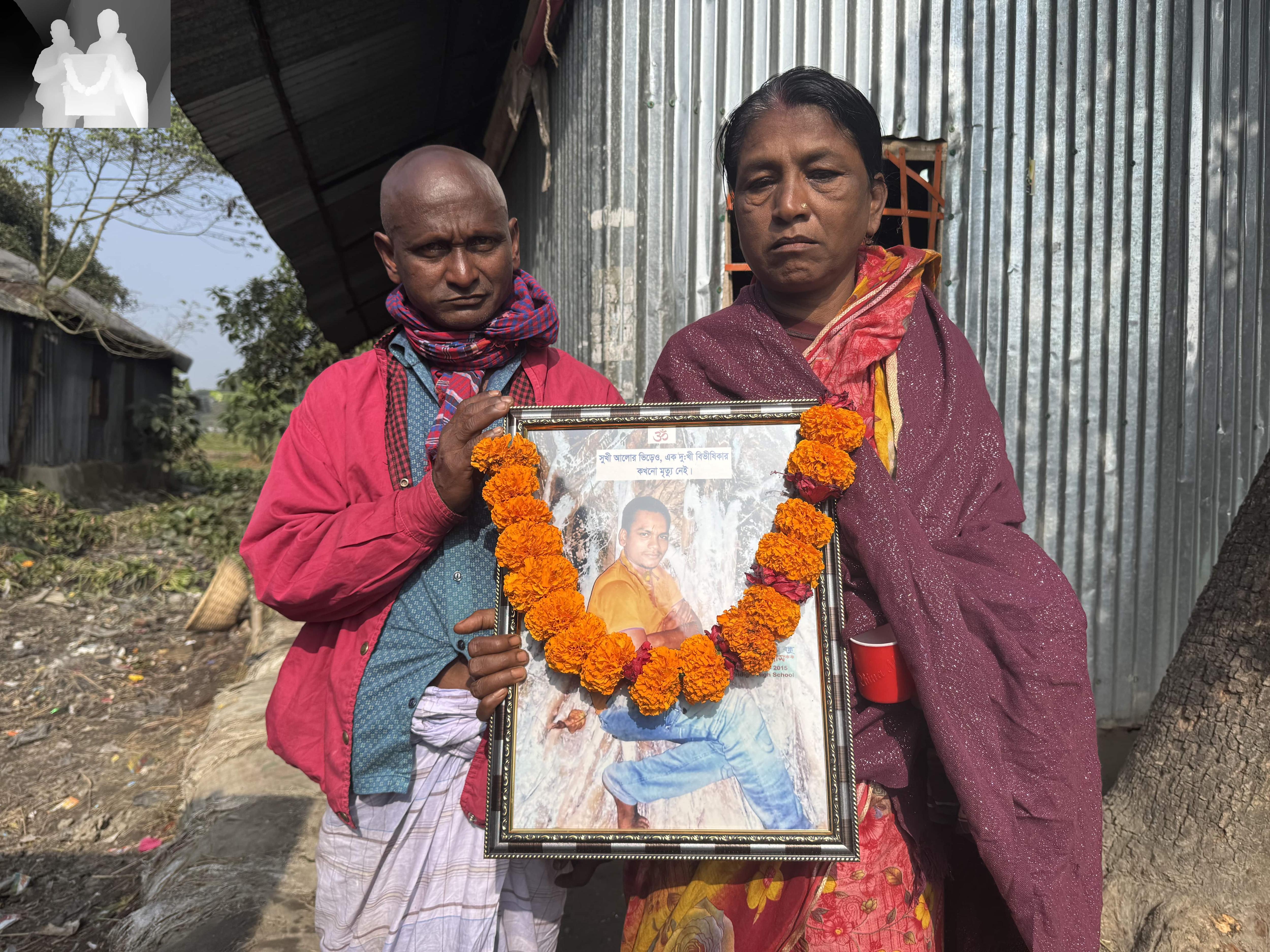 A south Asian man and woman, both old, very sad, stand in front of a metal shack, hold a photo of man, a garland across it.
