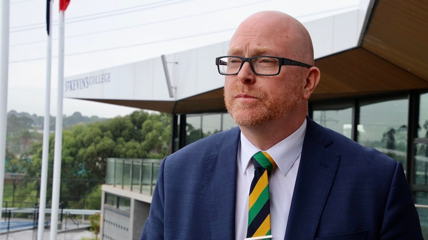 John Crowley, wearing glasses and a suit, looks at the camera while sitting behind a desk.