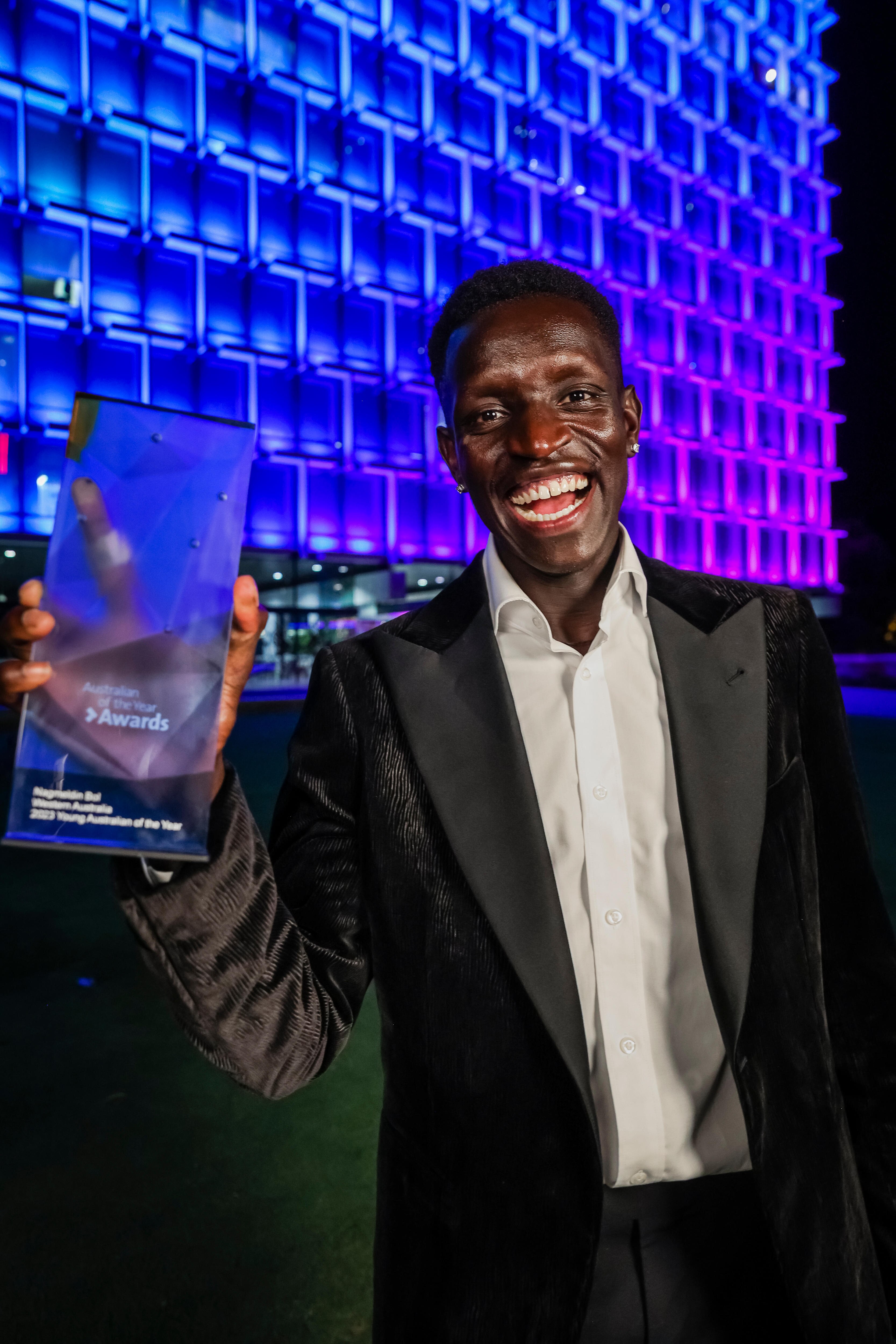 Athlete in suit smiles broadly whilst holding his australian of the year award in front of a stage 