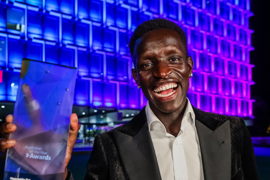 Athlete in suit smiles broadly whilst holding his australian of the year award in front of a stage 