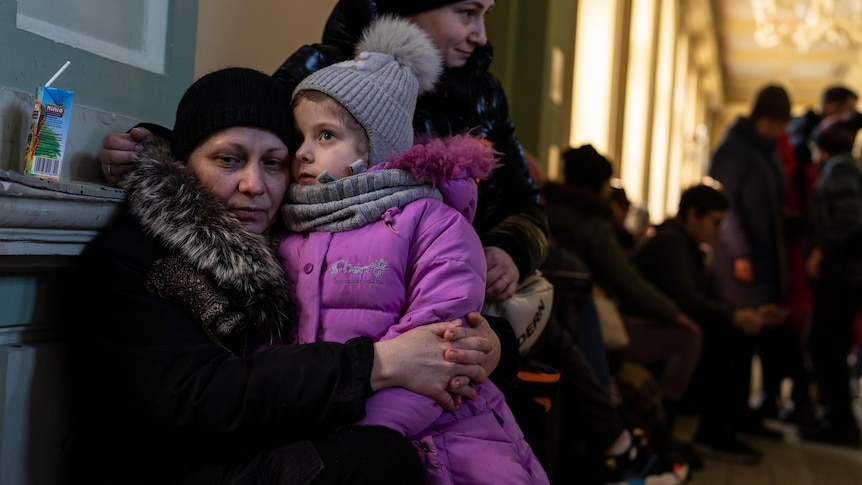 A woman in warm clothing sits on a seat on a train platform with a child sitting on her lap.