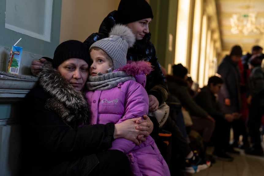 A woman in warm clothing sits on a seat on a train platform with a child sitting on her lap.