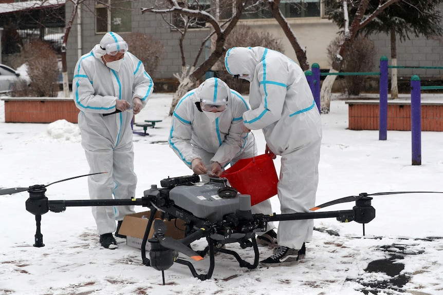 Three people in PPE stand outside a building in the snow preparing a large drone to disinfect a building.