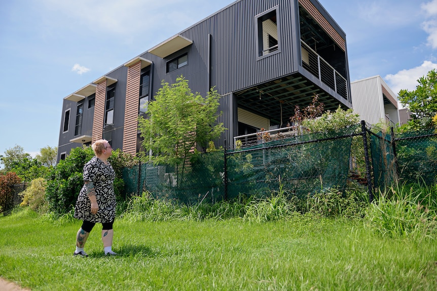 A woman stands outside a modular looking home, looking up at it, on a sunny day in the Top End.