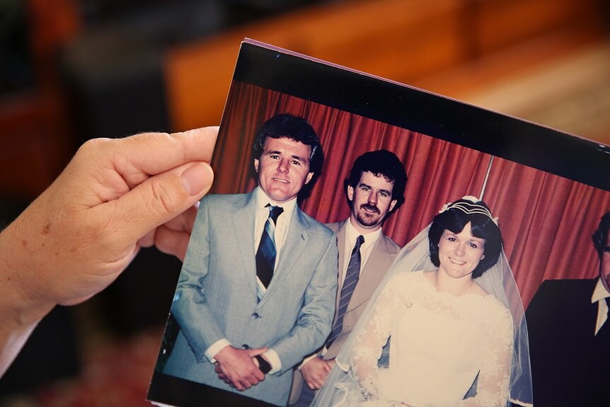 A woman's hand holds a photo of a wedding party from the 1980s