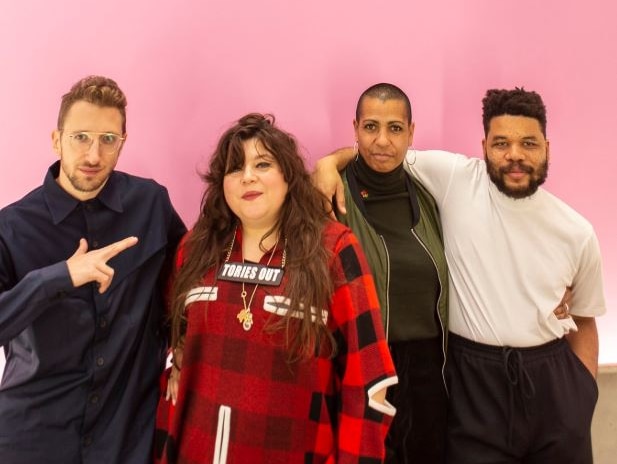 Four artists are arm-in-arm as they pose in front of a pale pink backdrop with the Turner Prize logo.