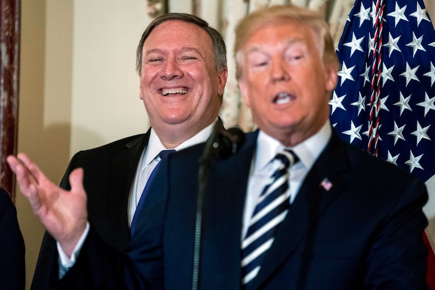 Mike Pompeo wears a big grin on his face as he stands behind Donald Trump, gesturing with his right hand, mid-speech