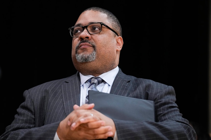 A close up of Manhattan District Attorney Alvin Bragg in a black pinstriped suit, holding a folder with a neutral expression