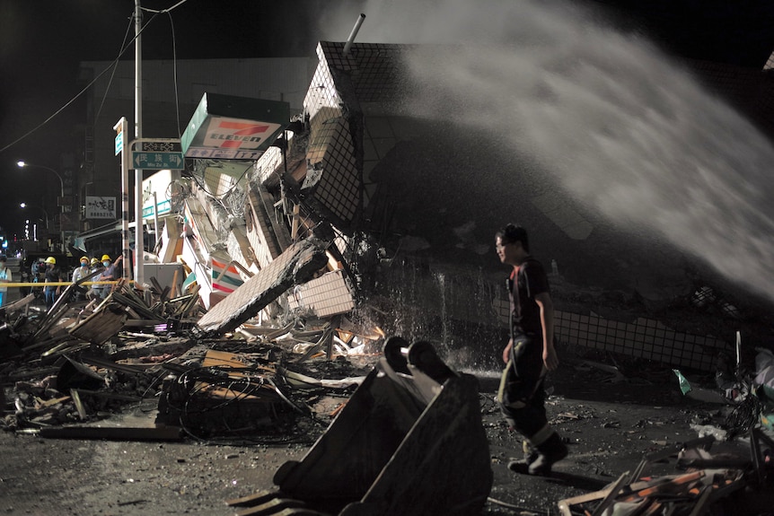 A person walks through rubble at night as a hose sprays water in an arc over their head. 