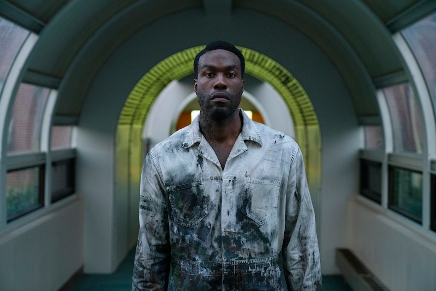 Yahya Abdul-Mateen II in a beaten-up and soiled white shirt lurches vacantly toward the camera, framed by a lime green archway.