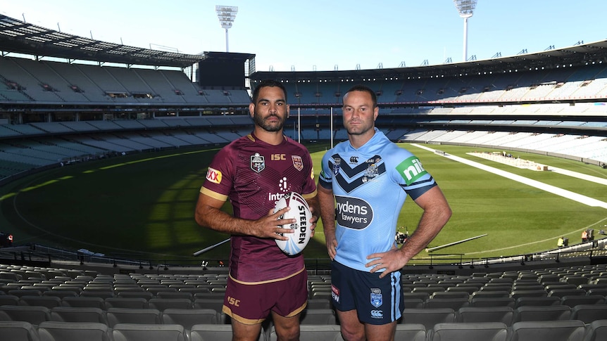 Greg Inglis holding a ball wearing a Maroons kit stands next to Boyd Cordner in a Blues kit with the MCG in the background.