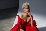 Montaigne wears a red dress and black shoes with fishnet socks. She bends down in front of white background and looks to camera.