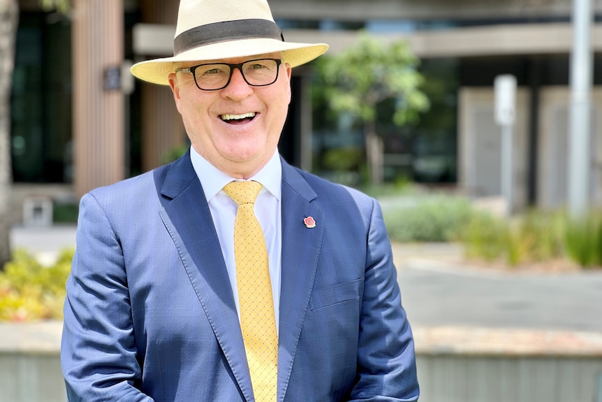 A man wearing glasses, a blue suit, yellow tie and yellow hat smiles at the camera.