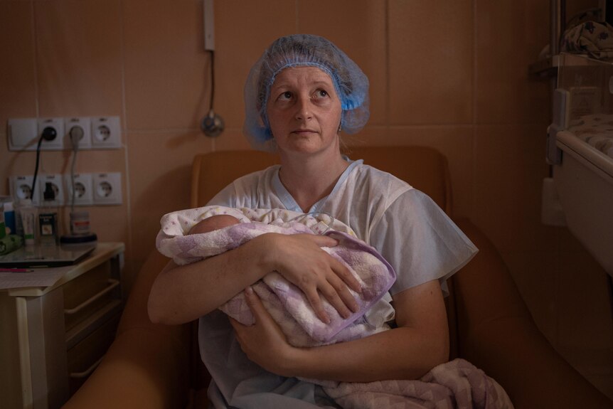 A woman in a blue hospital hair net clutches her baby to her chest and looks up with a concerned expression.