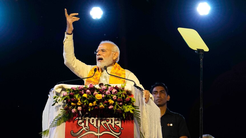 Modi stands at a podium decorated with flowers and speaks with a hand raised in the air. 
