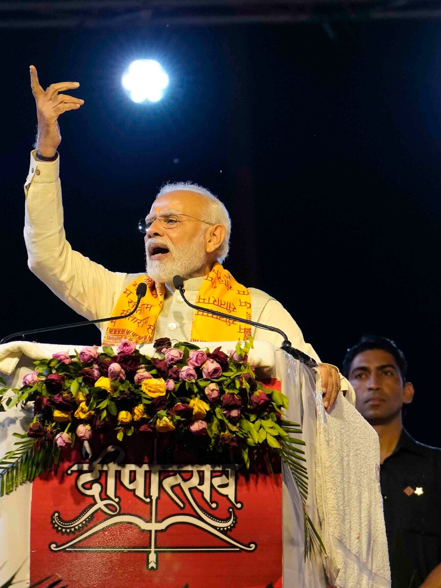 Modi stands at a podium decorated with flowers and speaks with a hand raised in the air. 