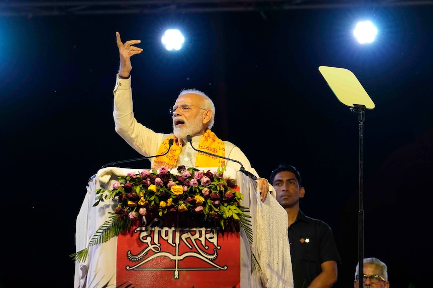 Modi stands at a podium decorated with flowers and speaks with a hand raised in the air. 