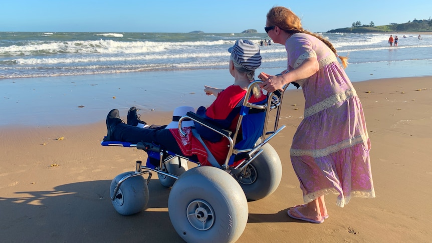 Mila in a beach wheelchair on the sand, facing the ocean, daughter Olga behind chair.