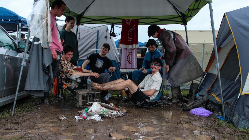 Several people either standing in mud or sitting on chairs with their feet up, smiling under a gazebo