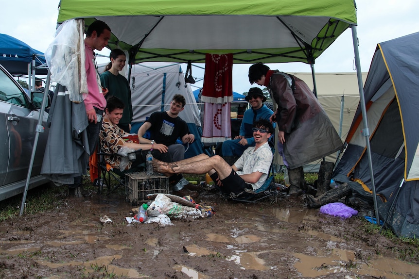 Several people either standing in mud or sitting on chairs with their feet up, smiling under a gazebo