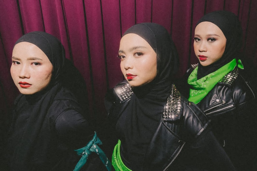 three women in a black hijab with bright green handkerchiefs accessories and metal stud jacket against a maroon curtain