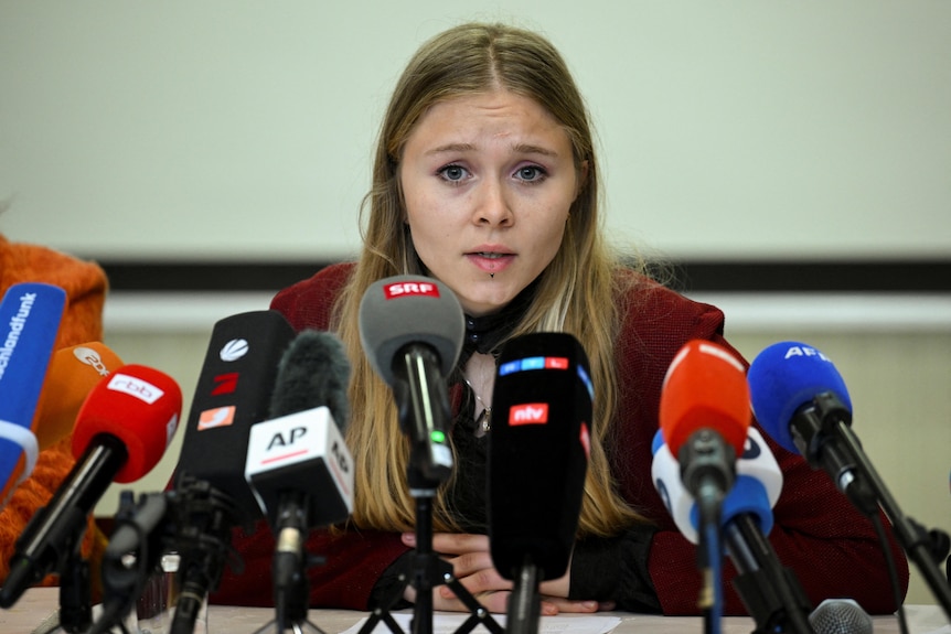 A young white woman with long blond hair speaking in front of a dozen microphones.