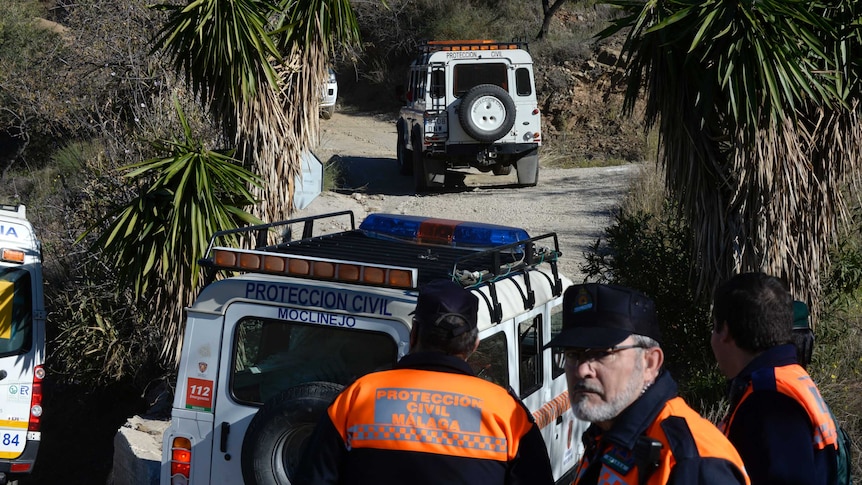 Three rescue workers stand in front of jeeps surrounded by palm trees.