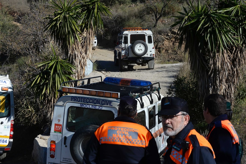 Three rescue workers stand in front of jeeps surrounded by palm trees.